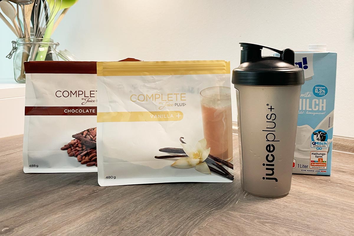 Zwei Packungen JuicePlus+ Complete Pulver, ein Shaker und eine Packung Milch auf einer Küchenarbeitsplatte.