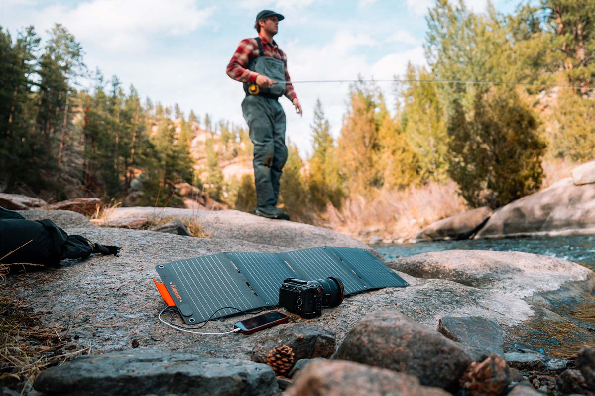 Ein Mann angelt in der Wildnis. Im Vordergrund liegt das Jackery SolarSaga 40 Watt Mini Solarpanel und läd eine Kamera und ein Smartphone auf.