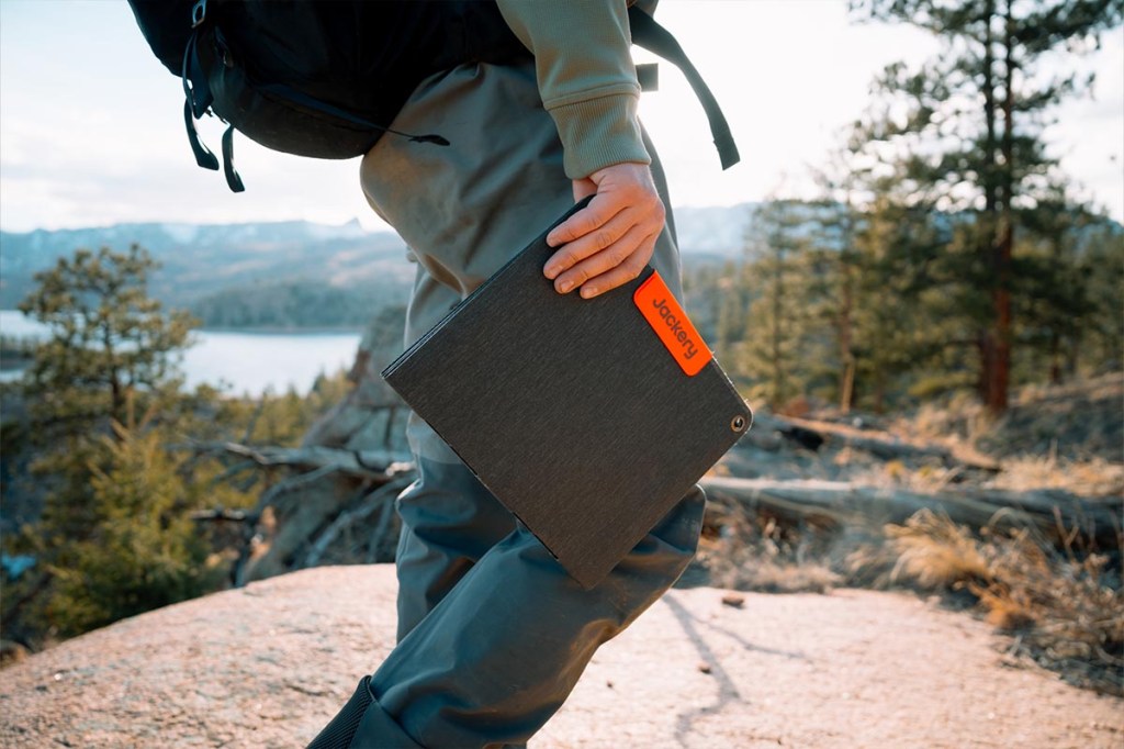 Eine Person mit Rucksack trägt in einer Hand das Jackery-Solarpanel in Natur-Umgebung.