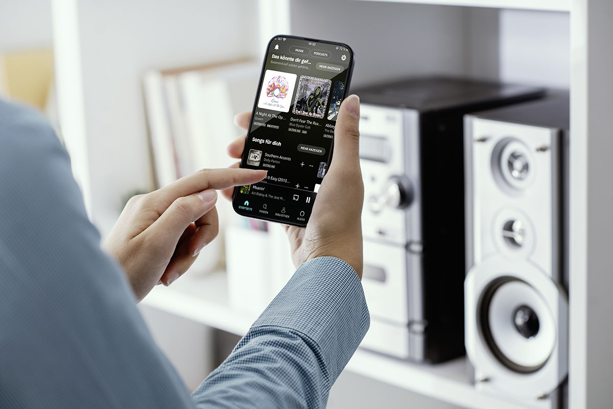 Man hält Smartphone vor Kompakt-Musik-Anlage in beiden Händen. Das Bild soll die Übertragung von Musik per Musikstreamingdienst vom Smartphone an eine kompatible Musikanlage zeigen.
