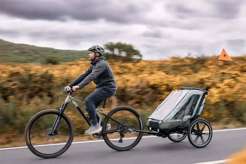 Mann fährt auf einem Fahrrad, er hat einen Kinderanhänger hinter sich, im Hintergrund Natur