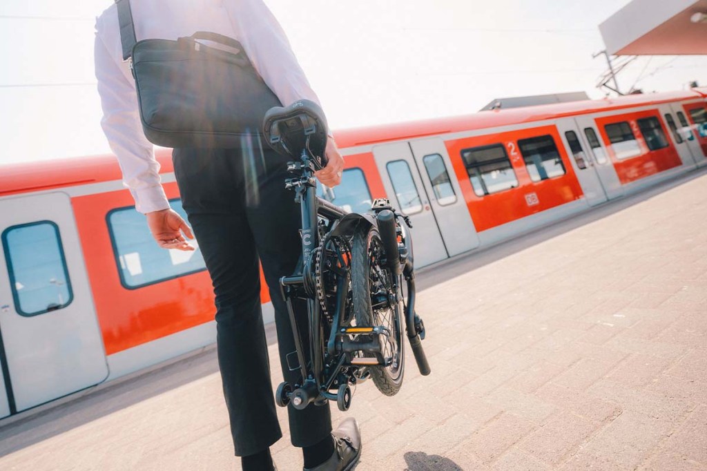 Person von hinten mit einem Faltrad in der Hand, steht am Bahnsteig, Bahn im Hintergrund