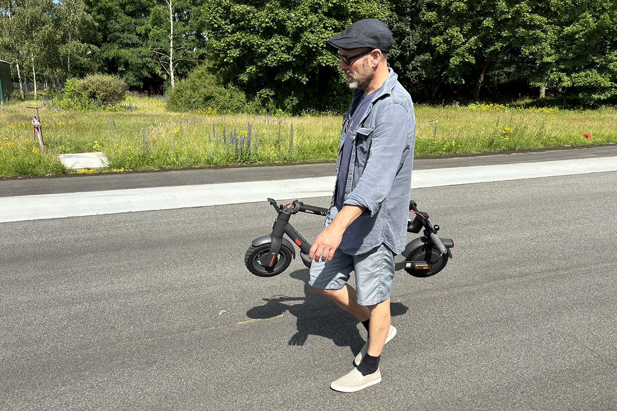 Mann trägt einen zusammengeklappten E-Scooter eine Straßen vor einer Rasenfläche entlang.