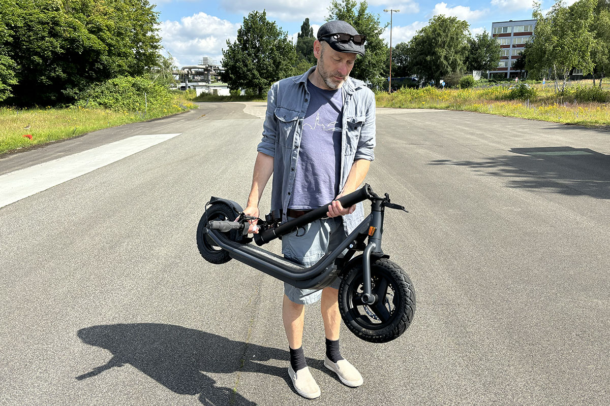 Mann trägt zusammengeklappten E-Scoooter und steht dabei auf einer Straße vor einem Waldstück.