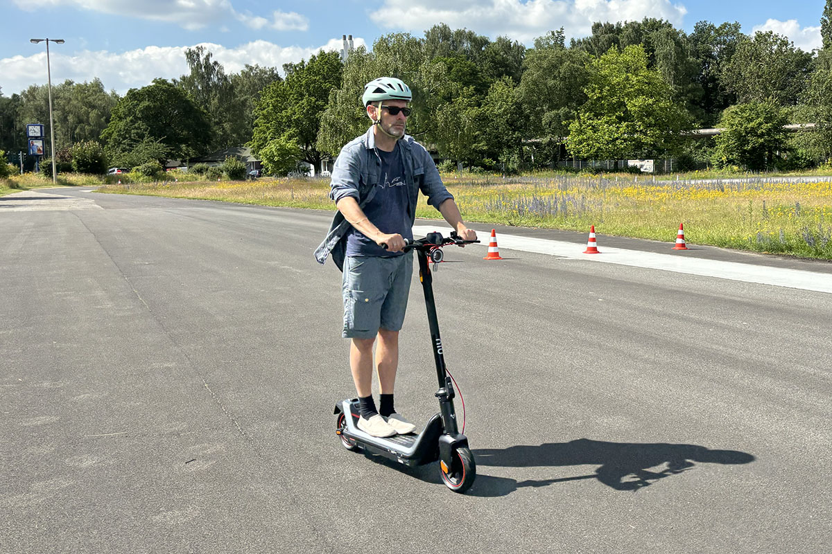 Mann fährt auf einem E-Scooter. Man sieht ihn von vorn an einem Waldstück vorbeifahrend.