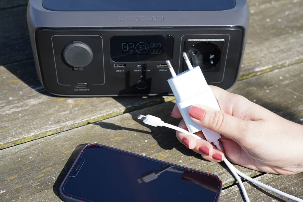 Eine Hand hält ein Ladekabel und ein Netzteil vor die River 3. Davor liegt ein Handy.