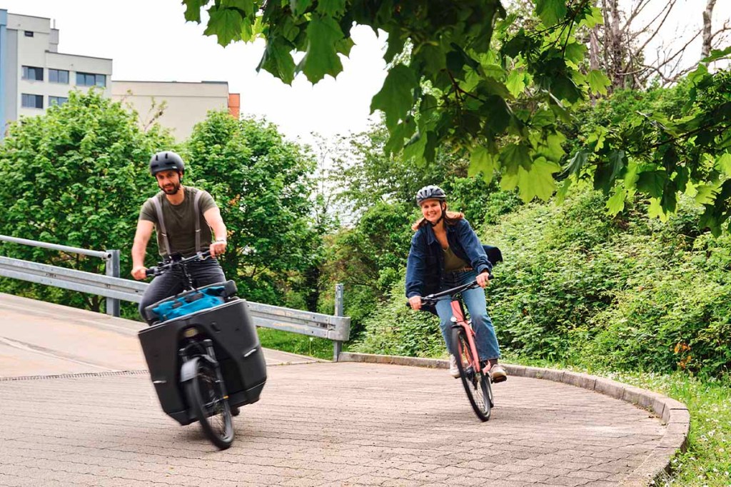 Ein Mann auf einem Lastenrad und eine Frau auf einem Fahrrad fahren auf einem breiten Fahradweg.