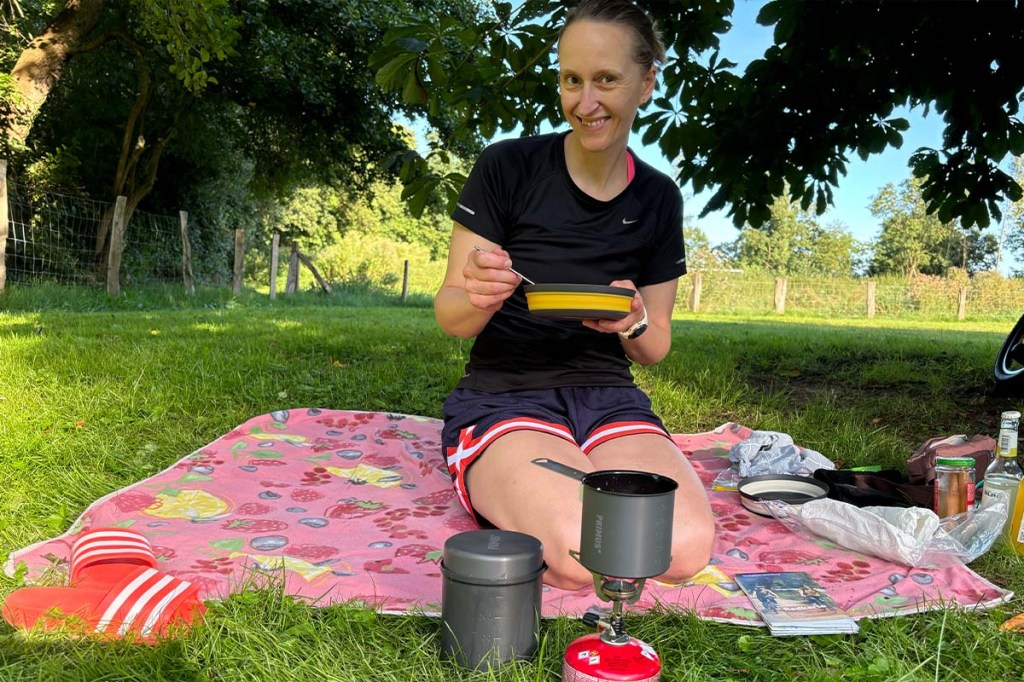 Frau auf einer Decke, hat Campinggeschirr in der Hand, Gaskocher im Vordergrund