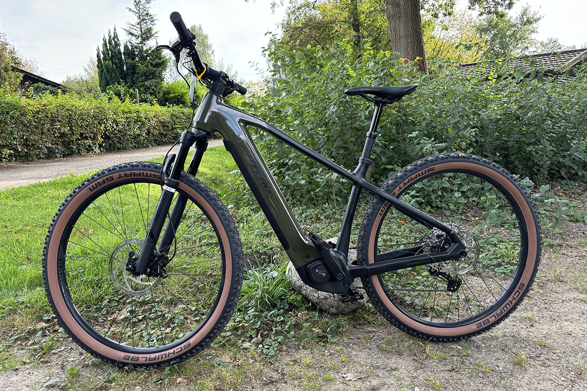 Sportliche Hardtail E-Mountainbike Stevens E-Agnello steht vor einer Hecke in einem Kleingartengebiet.