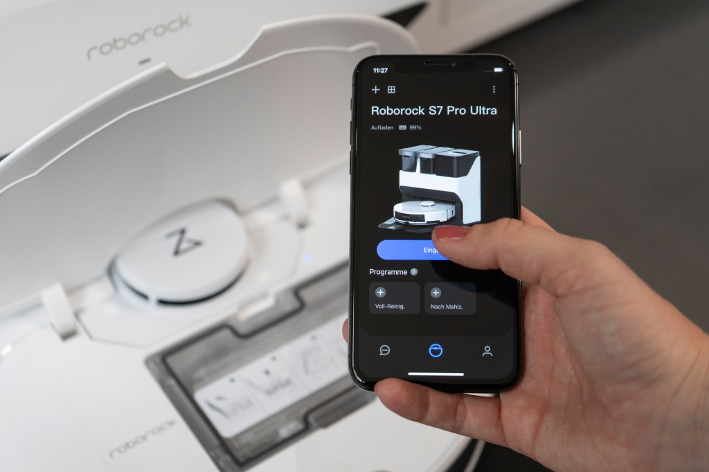 Eine Hand hält ein Smartphone mit geöffneter App eines Saugroboter vor einen Saugroboter.