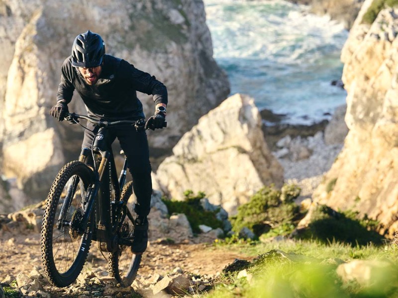 Mann fährt mit einem E-MTB einen steilen Trail hoch, Meer und Felsen im Hintergrund