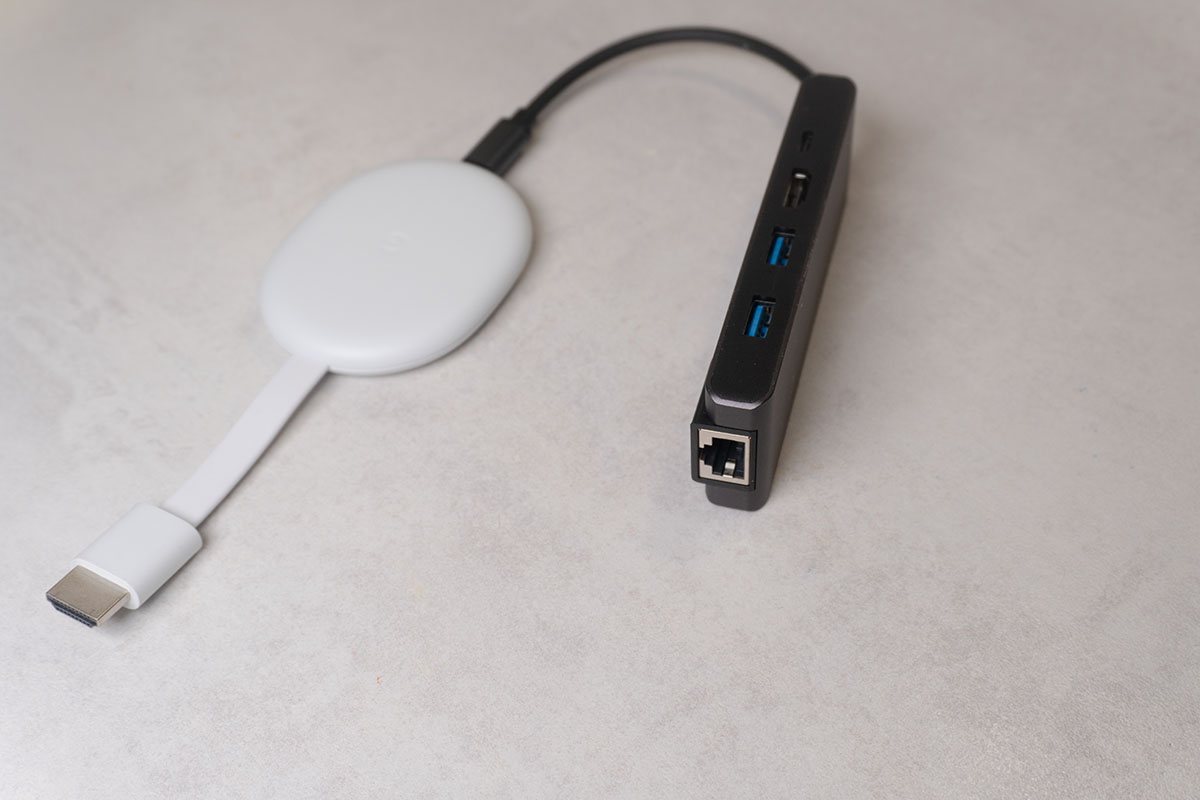 Weißer Streaming Stick Chromecast mit Google TV 4K ist via Kabel mit einem Adapter verbunden, der neben einem Netzwerkkabel-Steckplatz auch Anschlüsse für USB und HDMI bietet