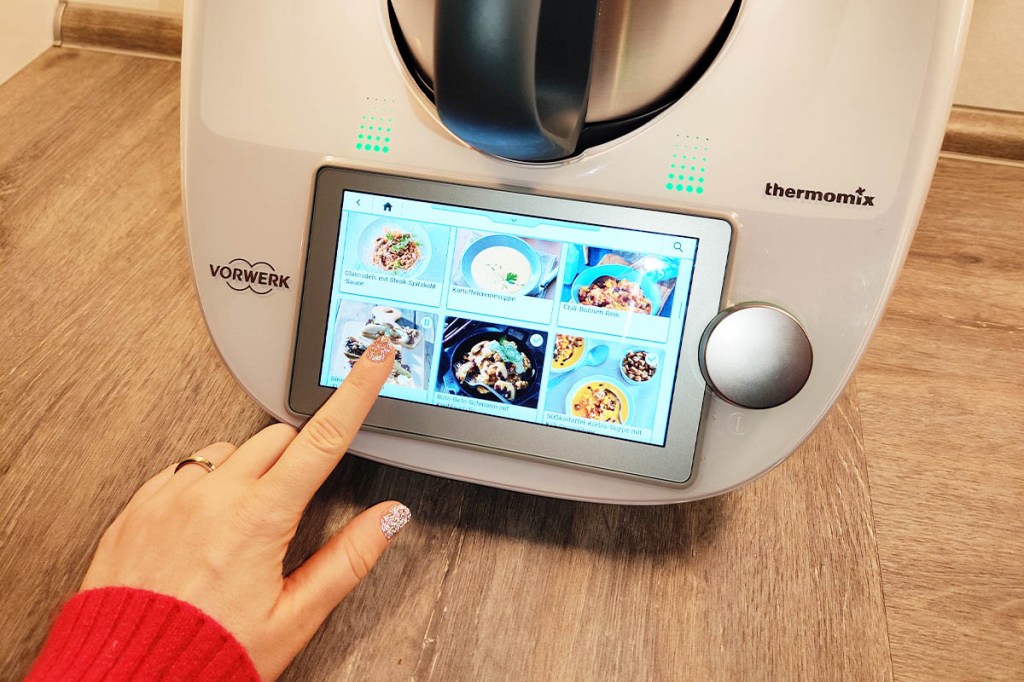 Eine Hand bedient das Display des Thermomix TM6, auf dem Rezepte zu sehen sind.