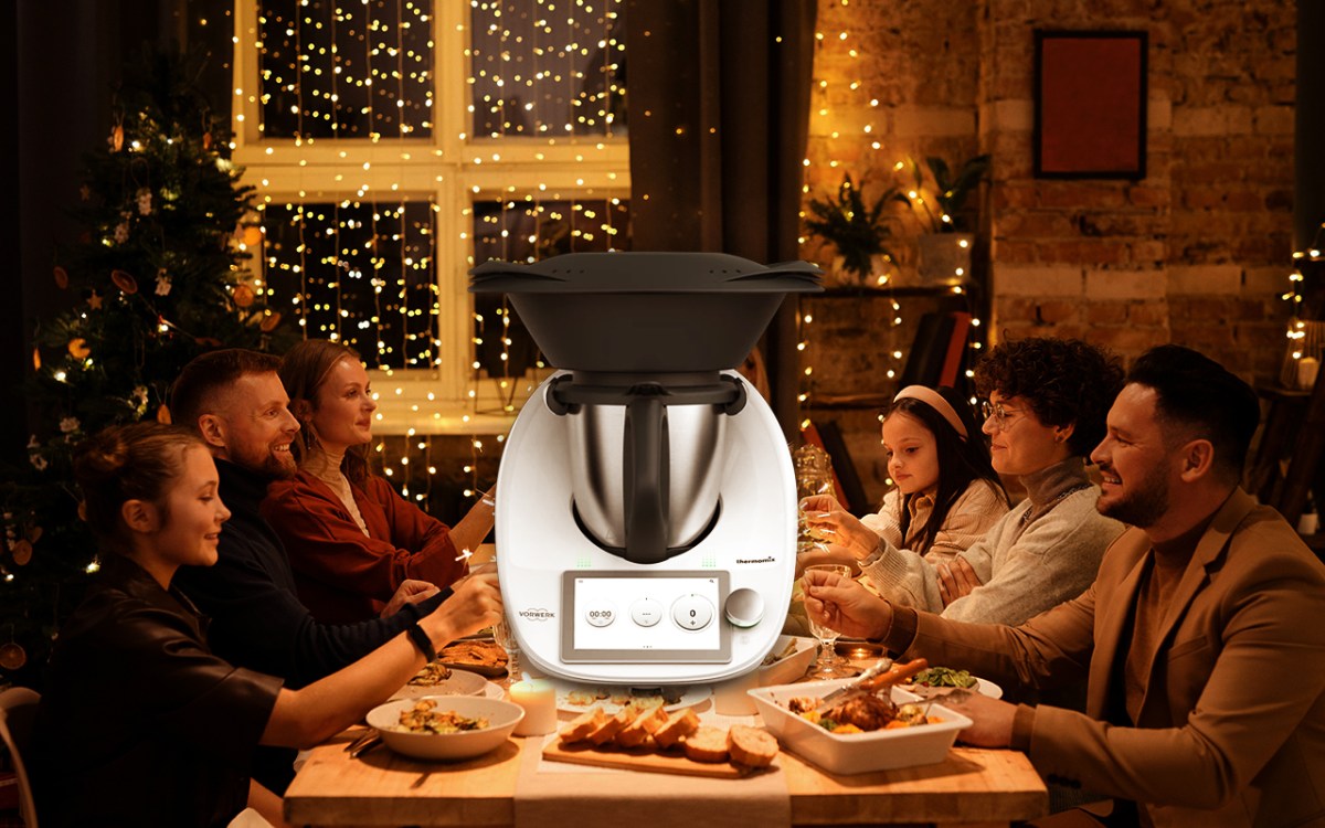 Eine Familie sitzt an einem Esstisch. Links ein Weihnachtsbaum, der Raum ist festlich geschmückt. Auf den Tisch ein Thermomix montiert.