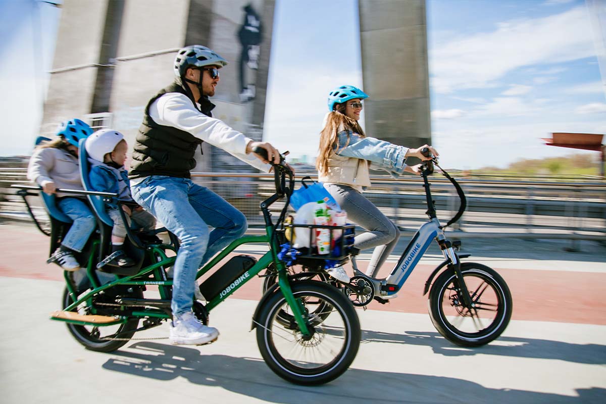 Zwei Personen fahren mit ihrem E-Bikes eine Straße entlang, die vordere Person hat zwei Kinder auf dem Rücksitz