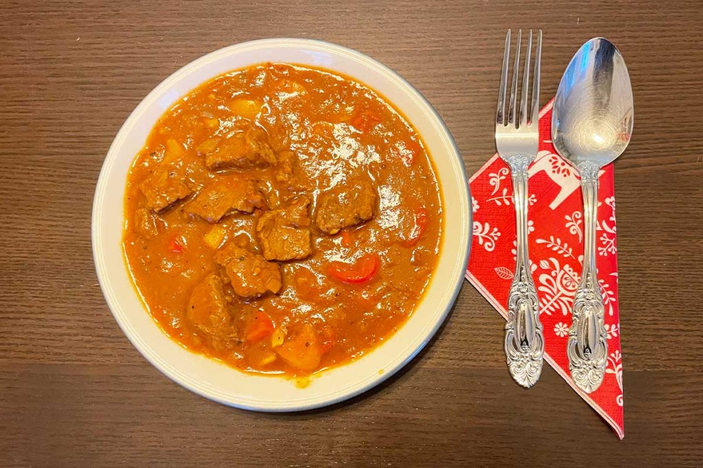 Ein Teller Gulasch von oben. Daneben liegen Gabel und Löffel mit Serviette auf dem Holztisch.