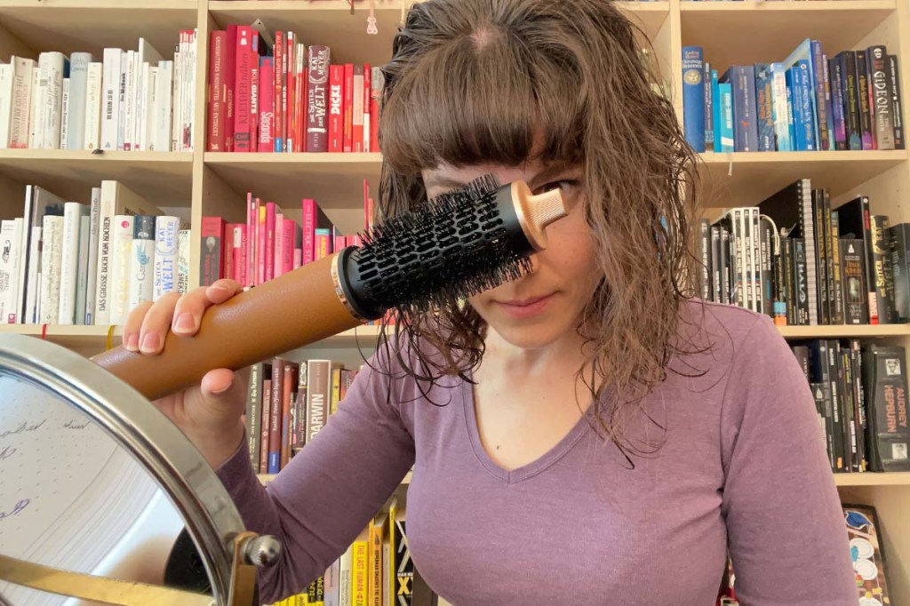 Eine Frau sitzt vor einem bunten Bücherregal und formt mit dem Dreame Airstyle Pro und dem Rundbürsten-Aufsatz den Pony.