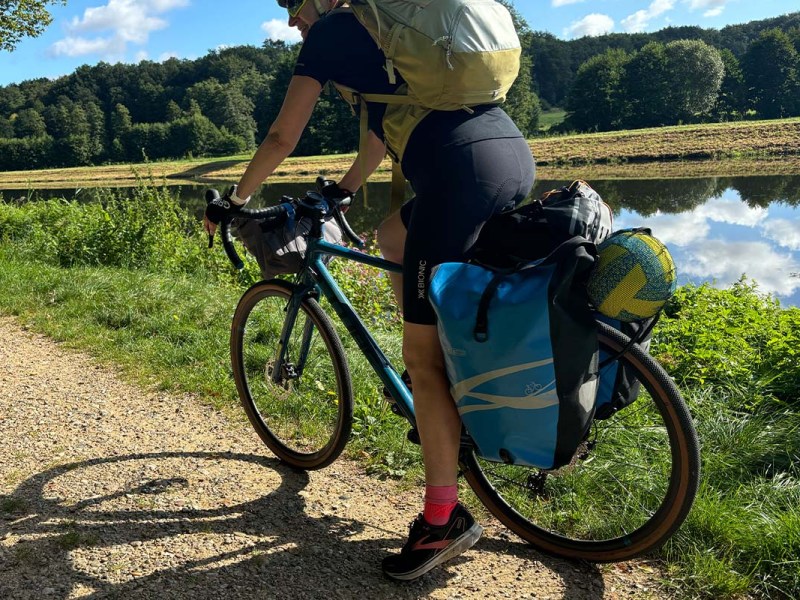 Anschnitt eines fahrradfahrers von hinten, der gepäckträger ist mit taschen voll beladen, ein kanal und natur im HIntergrund