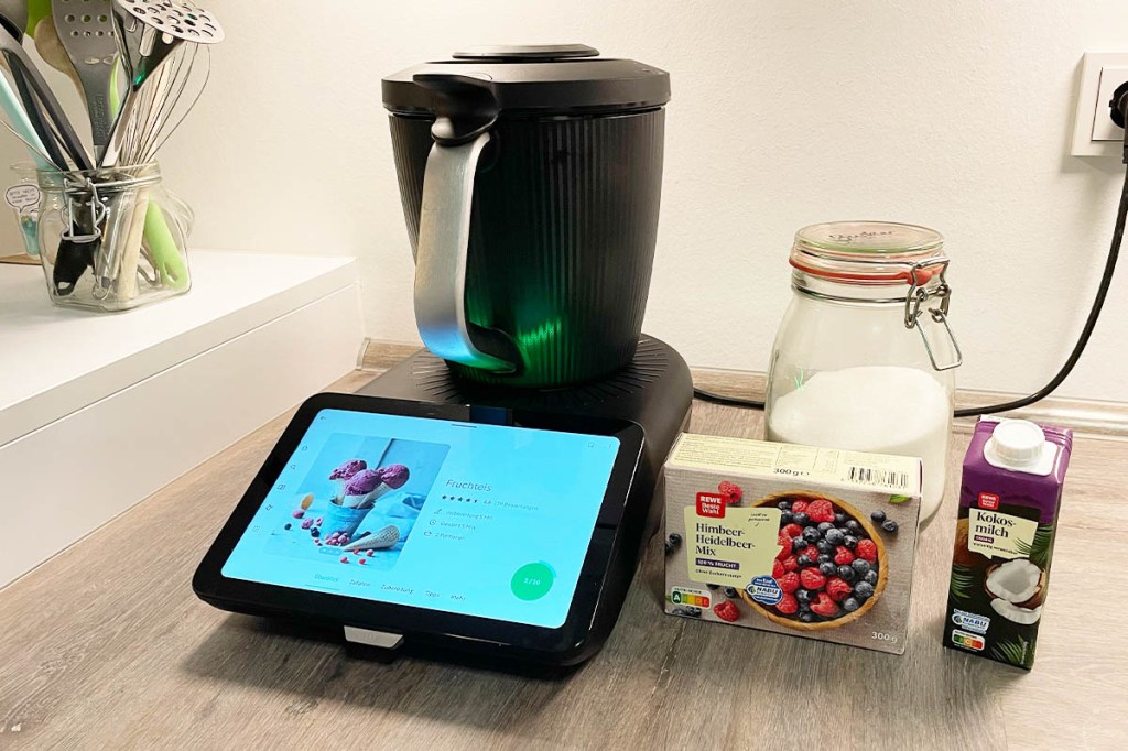 Der Thermomix TM7 mit leuchtendem Display neben Zutaten für Fruchteis auf einer Arbeitsplatte.